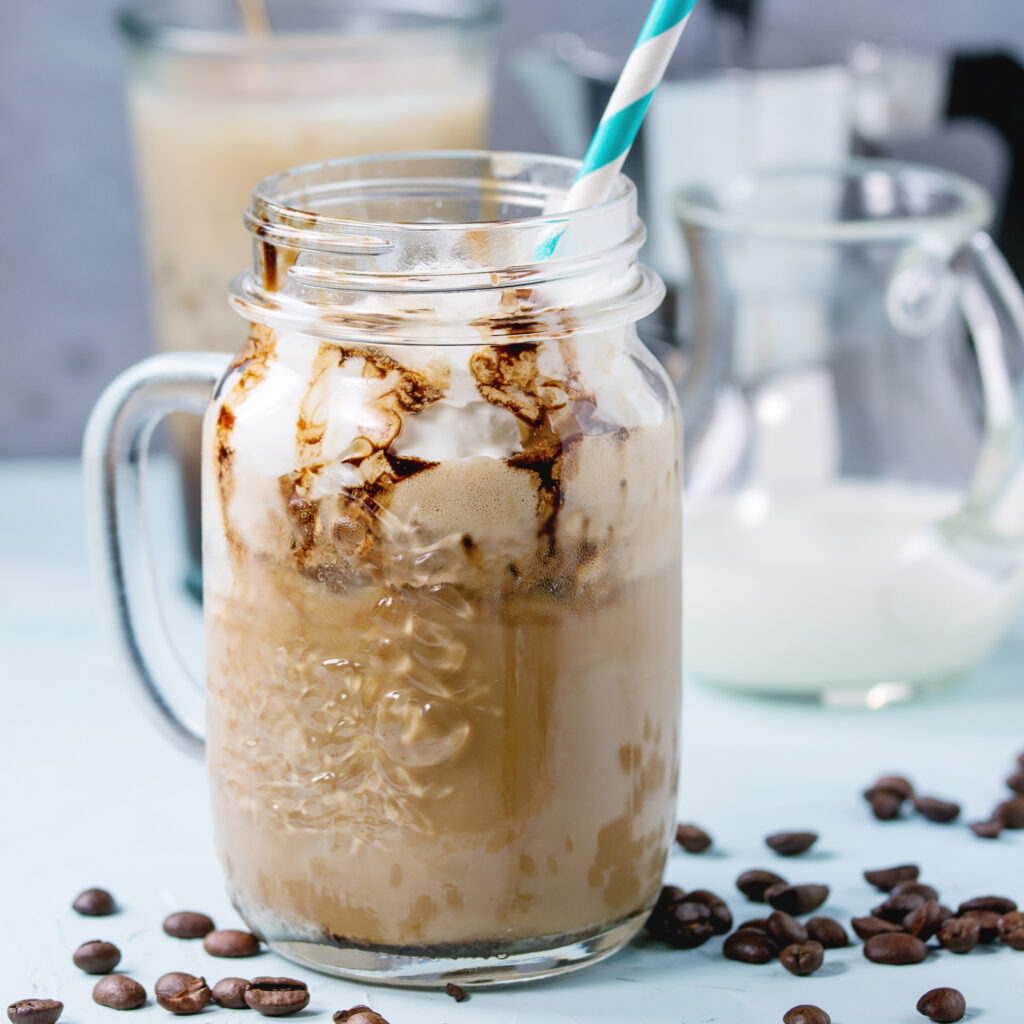 Glass mason jar with ice coffee with whipped cream, ice cream and chocolate sauce, served with coffee beans, coffe pot and jug of milk over light blue textured background. Square image