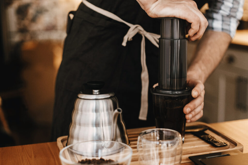 Professional barista preparing coffee by aeropress alternative method, brewing process. Hands on aeropress and glass cup, scales, manual grinder, coffee beans, kettle on wooden table